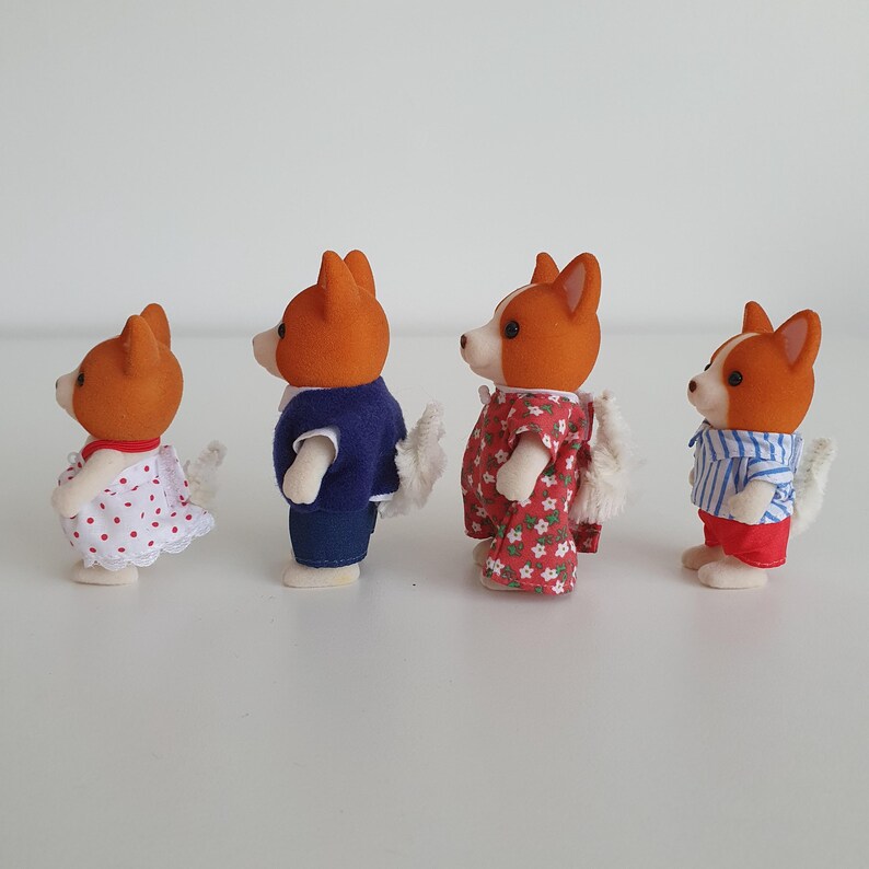 May include: Four miniature dog figurines with orange and white fur, each wearing different outfits. One wears a white dress with red polka dots, another a blue jacket and jeans, and the others wear floral and striped outfits.