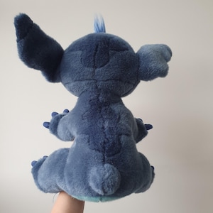May include: A plush toy of the character Stitch from the Disney movie Lilo & Stitch. The blue stuffed animal has large ears, a tuft of hair, and is in a seated position. The toy is made of soft, textured fabric.