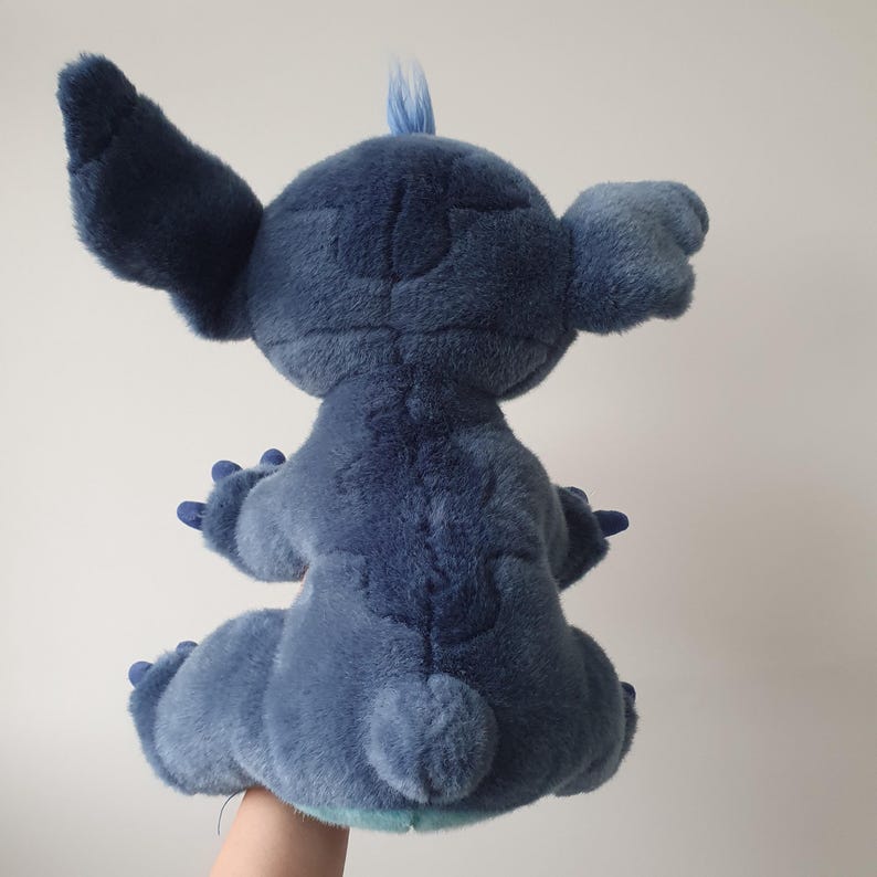 May include: A plush toy of the character Stitch from the Disney movie Lilo & Stitch. The blue stuffed animal has large ears, a tuft of hair, and is in a seated position. The toy is made of soft, textured fabric.
