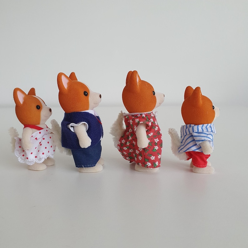 May include: Four toy corgi figures, each with orange and white fur, dressed in various outfits. One wears a white dress with red polka dots, another a navy blue suit, a third a red floral dress, and the last a striped shirt and red pants.