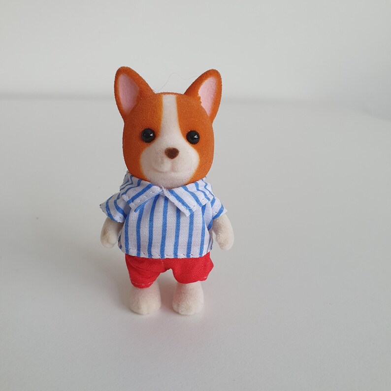 May include: A miniature dog figurine with a brown and white head, wearing a blue and white striped shirt and red shorts. The toy dog has black eyes and white paws, and is standing upright.