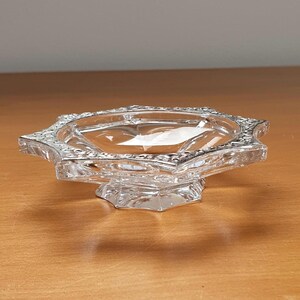 May include: A clear glass dish with a star-shaped rim and a pedestal base. The dish has a decorative, raised pattern around the rim.