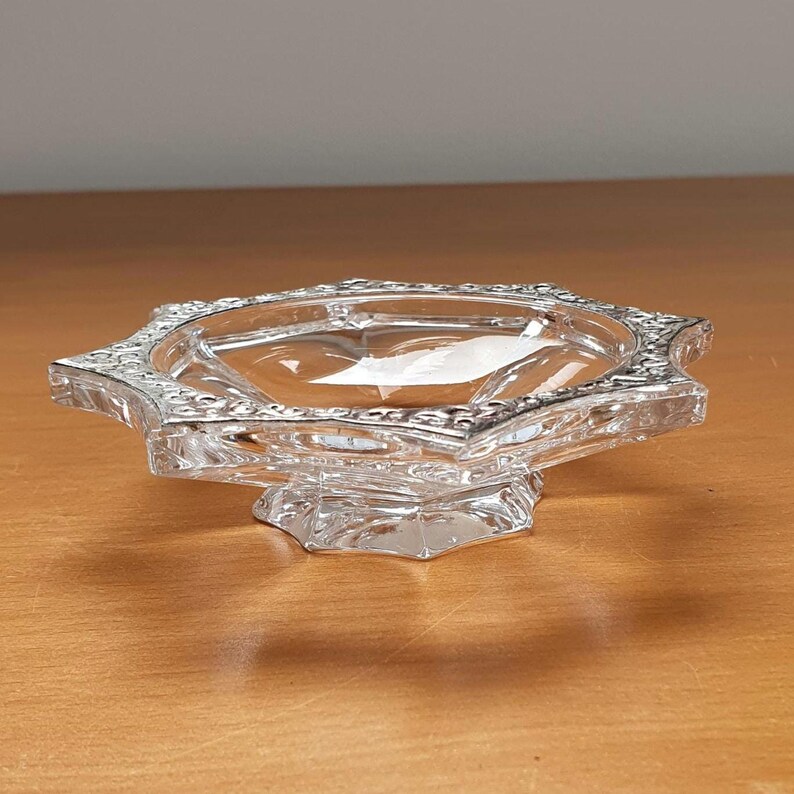 May include: A clear glass dish with a star-shaped rim and a pedestal base. The dish has a decorative, raised pattern around the rim.