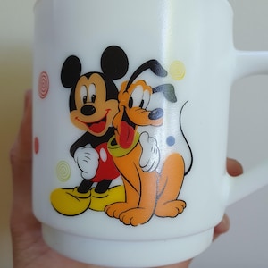 May include: White ceramic mug featuring Mickey Mouse and Pluto. Mickey is in black, red, yellow, and white, and Pluto is orange, yellow, and white. The mug has a handle and is decorated with colorful swirls and dots.