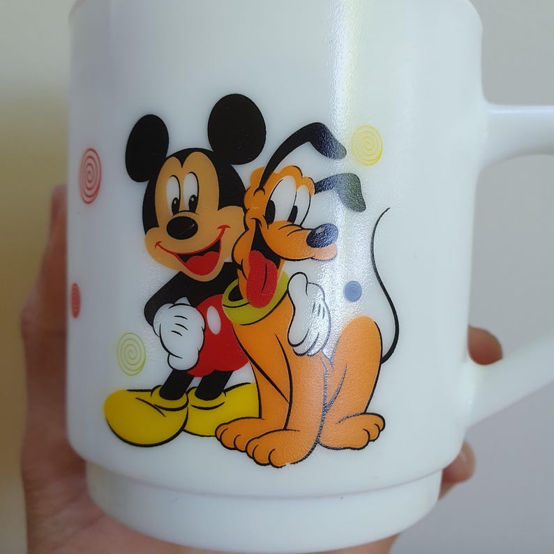 May include: White ceramic mug featuring Mickey Mouse and Pluto. Mickey is in black, red, yellow, and white, and Pluto is orange, yellow, and white. The mug has a handle and is decorated with colorful swirls and dots.