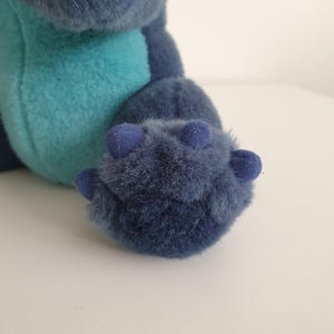 May include: Close-up of a plush toy's foot and leg. The foot is a deep blue with three small, rounded toes. The fur is soft and fluffy. The leg is a lighter blue-green color.