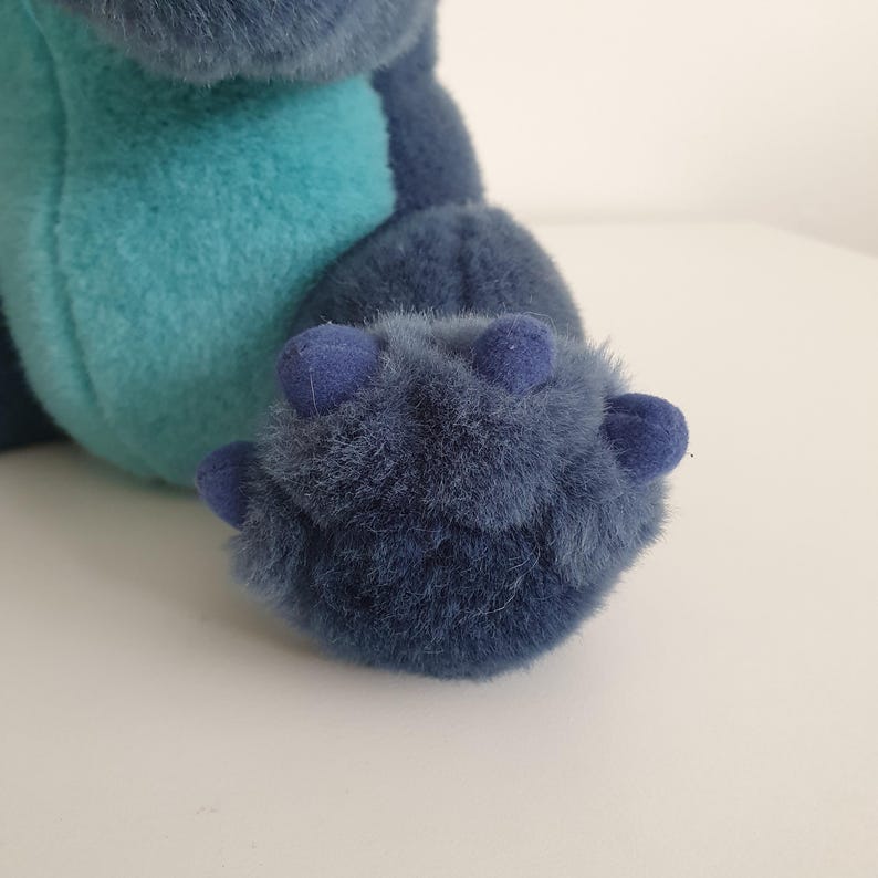 May include: Close-up of a plush toy's foot and leg. The foot is a deep blue with three small, rounded toes. The fur is soft and fluffy. The leg is a lighter blue-green color.