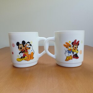 May include: Two white ceramic mugs with cartoon character designs. One mug features Mickey Mouse and Pluto, the other features Minnie Mouse and Daisy Duck. Each mug has a white handle and colorful accents.