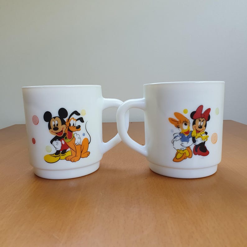 May include: Two white ceramic mugs with cartoon character designs. One mug features Mickey Mouse and Pluto, the other features Minnie Mouse and Daisy Duck. Each mug has a white handle and colorful accents.