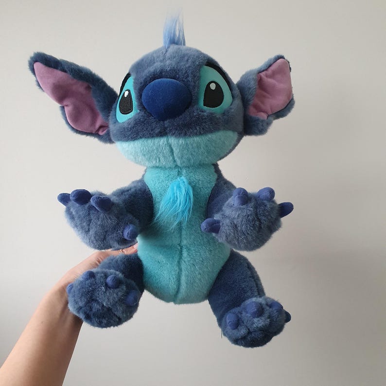 May include: A plush toy of the character Stitch from the animated film Lilo & Stitch. The toy is primarily blue with lighter blue and pink accents. It has large ears, a tuft of hair, and a fluffy chest. The toy is being held up.