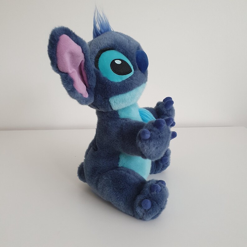 May include: A plush toy of the character Stitch from the animated film Lilo & Stitch. The toy is primarily blue with lighter blue and pink accents. It has large ears, big eyes, and is in a sitting position.