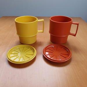 Könnte beinhalten: Zwei stapelbare Plastikbecher mit Deckeln, einer gelb und einer orange. Die Becher haben ein schlichtes Design mit einem Ring in der Mitte.