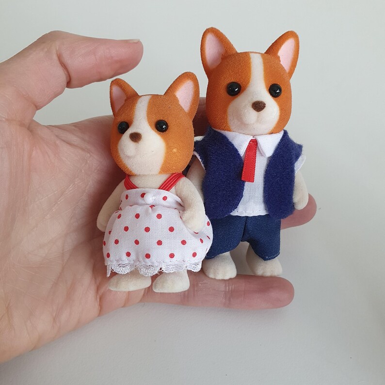 May include: Two miniature dog figurines. One wears a white dress with red polka dots and red straps. The other wears a blue vest, white shirt, red tie, and blue pants. Both have brown and white fur.
