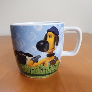 以下が含まれることがあります： 青い帽子をかぶり、笛を持っている犬の漫画のキャラクターが描かれた白いセラミックマグ。マグカップには、緑色のニットの縁と、漫画のグラフィックが描かれた水色の背景があります。ハンドルは白です。