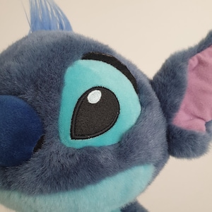May include: Close-up of a plush Stitch toy. The toy features a blue and turquoise color scheme with a tuft of blue hair. The large eyes have black outlines and a white highlight. The ears are a mix of blue and pink.