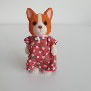 May include: A toy corgi dog figure with orange and white fur, wearing a red dress with a white floral pattern. The dress has short sleeves and a white collar. The figure is standing on a white surface.