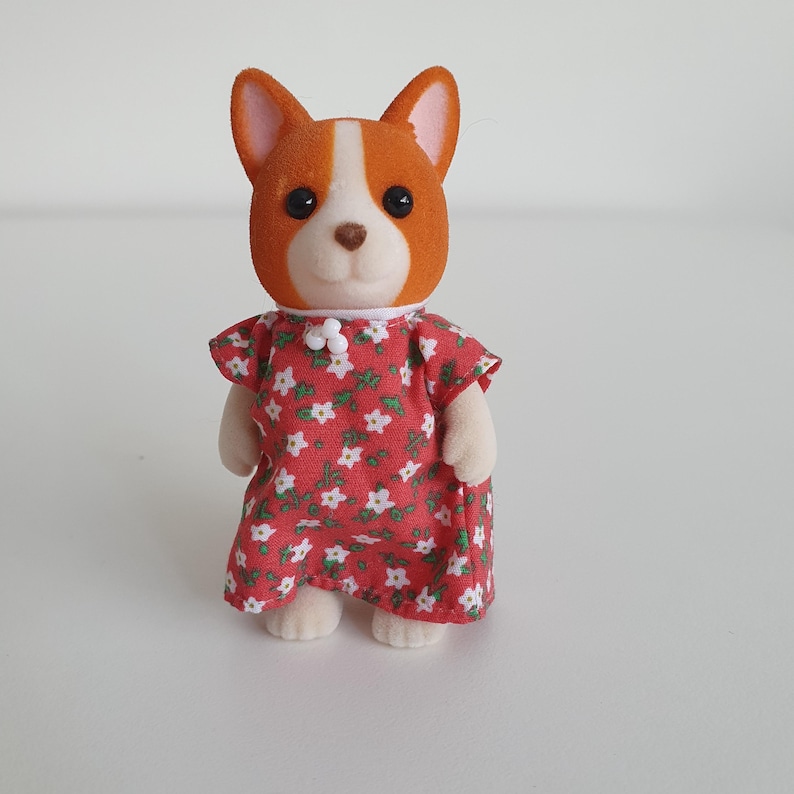 May include: A toy corgi dog figure with orange and white fur, wearing a red dress with a white floral pattern. The dress has short sleeves and a white collar. The figure is standing on a white surface.
