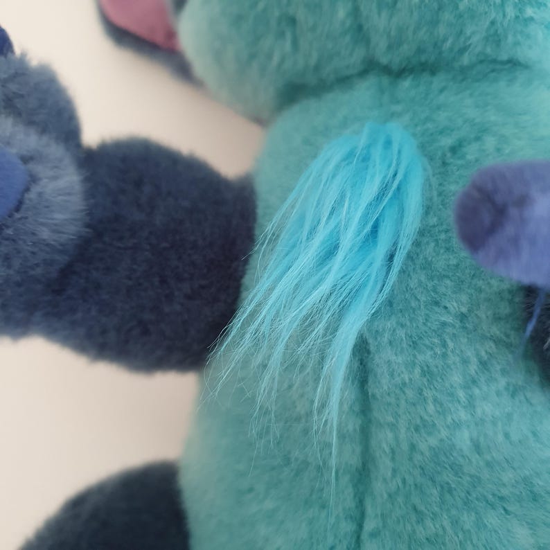 May include: A plush toy in shades of blue and teal, with a tuft of light blue fur. The toy has a soft, fuzzy texture and is likely a character from a popular animated film. The plushie's ear is a muted purple.