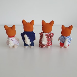 May include: Four toy fox figures with orange heads and white fluffy tails. The figures are dressed in various outfits: a white dress with red polka dots, a blue jacket and jeans, a red floral dress, and a blue and white striped shirt with red pants.
