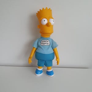 Op de afbeelding: Een Bart Simpson-pop, met een gele kop en stekelig haar, grote ogen en een prominente neus. De pop draagt een lichtblauw t-shirt met de tekst "WHOA MAMA!" en een blauwe korte broek. Blauwe schoenen met witte zolen maken de look compleet.