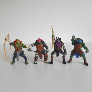 May include: Four Teenage Mutant Ninja Turtles action figures standing in a row. The first figure wears an orange mask and holds nunchucks. The second wears a red mask and holds two sais. The third wears a purple mask and holds a bo staff. The fourth wears a blue mask and holds two swords.