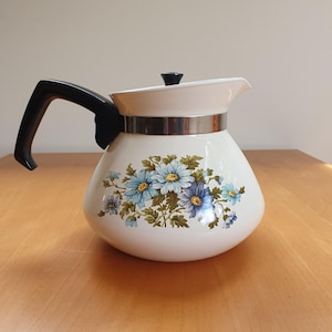 Könnte beinhalten: Weiße Keramik-Teekanne mit schwarzem Griff und Deckel. Die Teekanne hat ein Blumendesign mit blauen und gelben Blumen und grünen Blättern. Ein silbernes Band umschließt die Mitte der Teekanne.