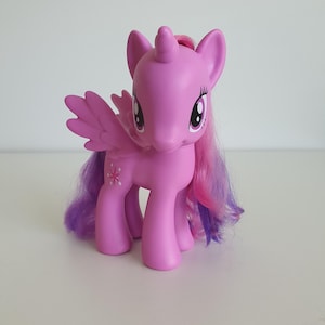 Puede incluir: Una figura de poni de plástico morado con un cuerno y alas. La figura tiene grandes ojos morados, una larga melena y cola con mechones de pelo rosa y morado, y una marca en forma de estrella en el costado. El poni se sostiene sobre cuatro patas.