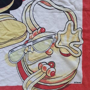 May include: A white and red towel featuring a cartoon handbag, sunglasses, and a star-shaped charm. The handbag has a yellow handle and red accents. The sunglasses have yellow lenses. The towel has a red border and the Disney logo.