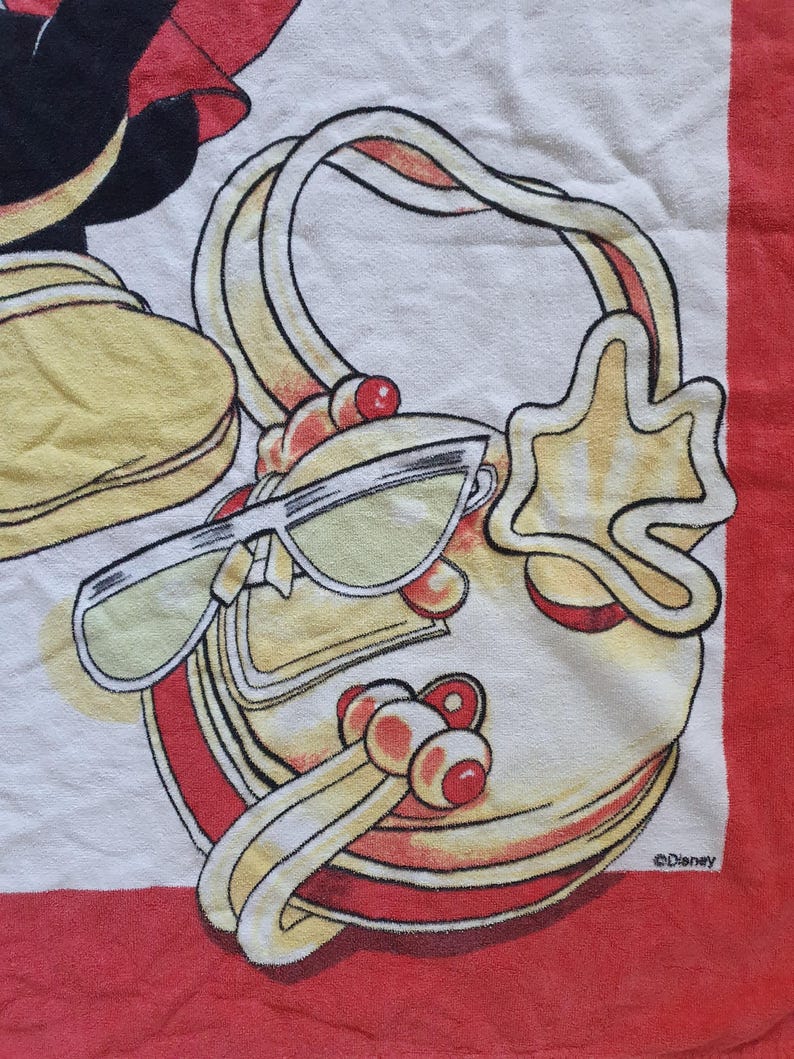 May include: A white and red towel featuring a cartoon handbag, sunglasses, and a star-shaped charm. The handbag has a yellow handle and red accents. The sunglasses have yellow lenses. The towel has a red border and the Disney logo.