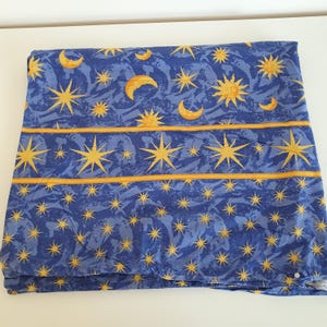May include: A folded blue fabric with a celestial pattern of yellow stars, moons, and suns. The design includes horizontal yellow stripes. The fabric has a textured appearance.