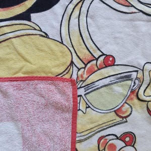 May include: A white and red striped beach towel with a red border. The towel features a cartoon illustration with yellow, black, and red accents, including sunglasses and a stylized design.