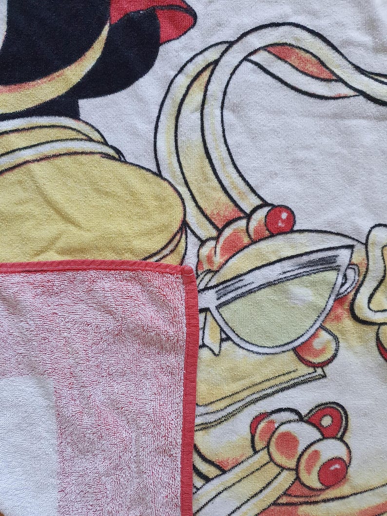 May include: A white and red striped beach towel with a red border. The towel features a cartoon illustration with yellow, black, and red accents, including sunglasses and a stylized design.