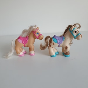 May include: Two plastic toy horses. The left horse is tan with a white mane and tail, a pink bridle, and a pink saddle. The right horse is cream with brown spots, a brown mane and tail, a blue bridle, and a purple saddle with blue hooves.