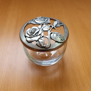 May include: A clear glass jar with a decorative metal lid featuring a rose design. The lid is silver-colored and has a circular shape with cut-out details. The jar sits on a wooden surface.