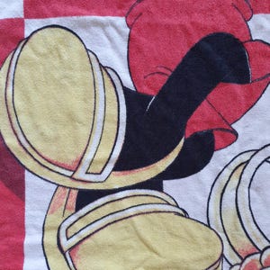May include: A white, red, and black cartoon character towel. The towel features a cartoon character's yellow shoes with white trim, red shorts, and black arms. The background has a red and white checkered pattern.
