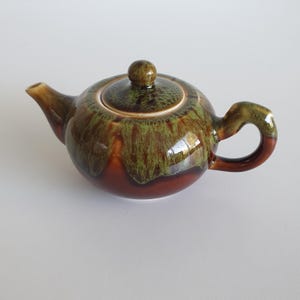 May include: A ceramic teapot featuring a glossy glaze with a drip pattern in shades of moss green and deep chocolate brown. The teapot has a rounded body, a curved handle, a short spout, and a matching lid with a small knob handle on top.