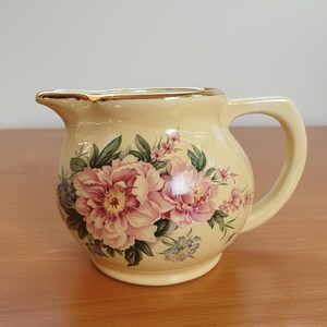 May include: A cream-colored ceramic creamer with a gold rim and a floral design featuring pink and blue flowers.