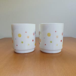 May include: Two white ceramic drinking glasses with a colorful polka dot and swirl design. The dots are red, yellow, green, and orange. The glasses are cylindrical with a slightly flared base, and are sitting on a wooden surface.