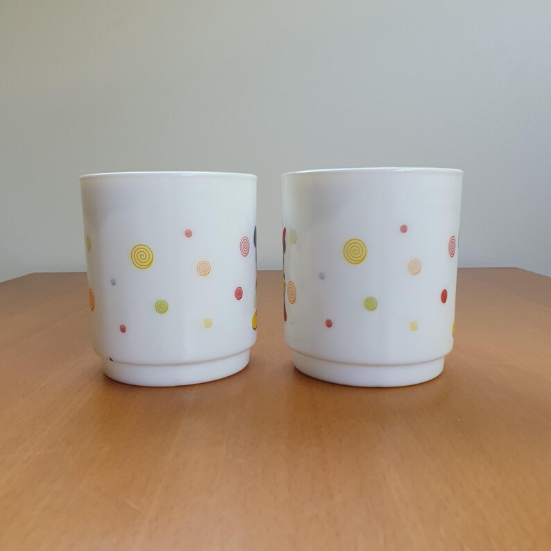 May include: Two white ceramic drinking glasses with a colorful polka dot and swirl design. The dots are red, yellow, green, and orange. The glasses are cylindrical with a slightly flared base, and are sitting on a wooden surface.