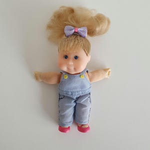 Puede incluir: Una muñeca pequeña con cabello rubio recogido en una coleta alta, sujeta con un lazo morado claro de lunares. La muñeca tiene cara redonda, ojos azules y viste un peto de mezclilla azul claro con botones amarillos sobre una camisa color melocotón.