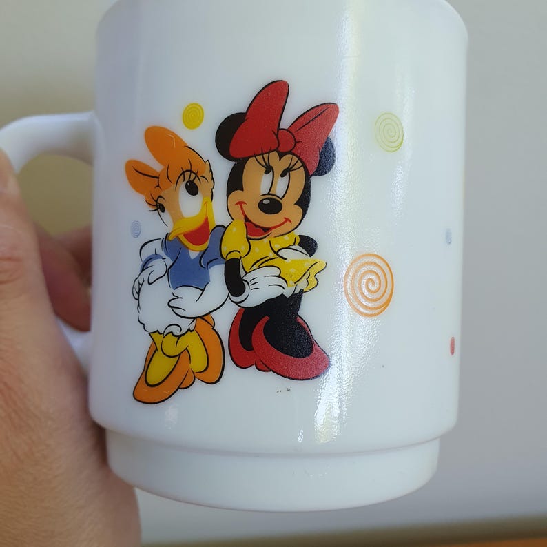 May include: White ceramic mug featuring Minnie Mouse and Daisy Duck. Minnie Mouse is wearing a red bow and yellow dress, while Daisy Duck is in a blue top and orange bow. The mug has a handle and is decorated with colorful polka dots and swirls.