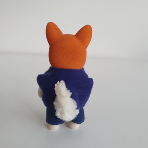 May include: A small orange fox figurine wearing a dark blue jacket and blue jeans. The fox has a fluffy white tail and is standing on a white surface. The figurine is a toy.
