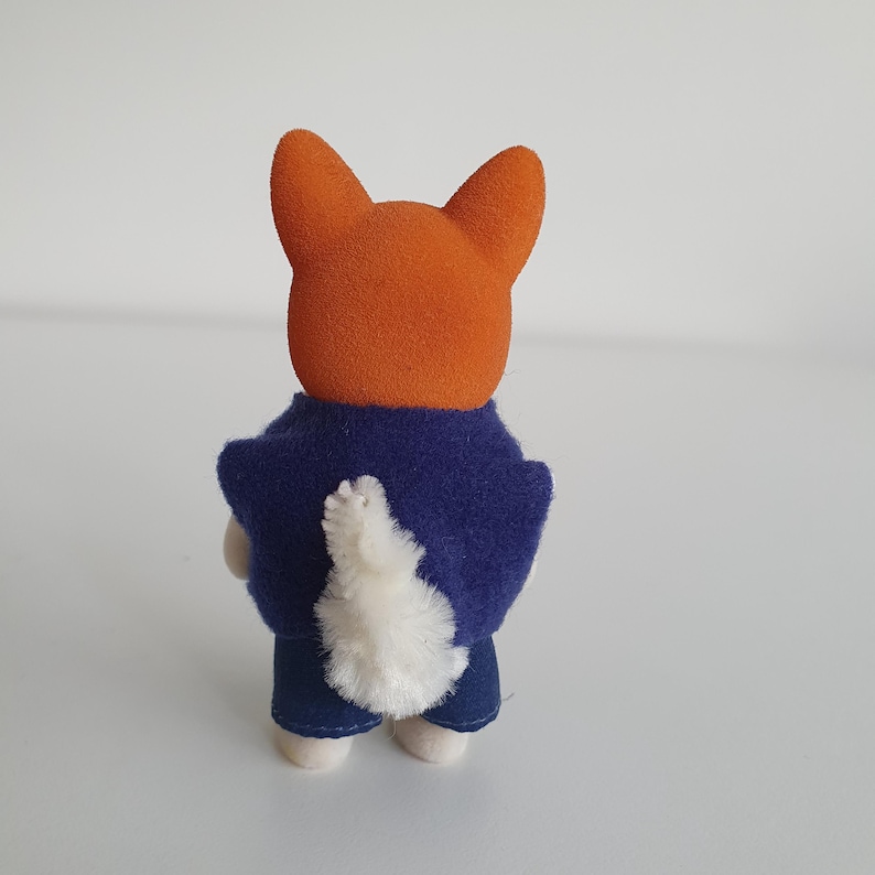 May include: A small orange fox figurine wearing a dark blue jacket and blue jeans. The fox has a fluffy white tail and is standing on a white surface. The figurine is a toy.