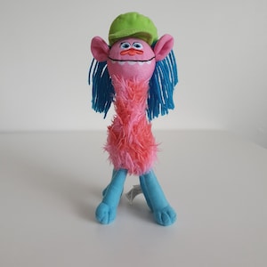 May include: A plush toy character with a pink face, large blue eyes, and orange eyebrows. It has long blue yarn hair, a lime green cap, and a fuzzy pink torso. The lower body consists of two light blue legs with visible stitched feet.