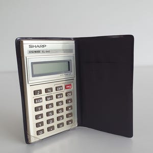Puede incluir: Una calculadora Sharp ELSI MATE EL-842 vintage en una funda negra. La calculadora tiene una cara plateada con teclas en blanco y negro, una pantalla digital y el texto "SHARP ELSI MATE EL-842". La funda está abierta, revelando la calculadora.