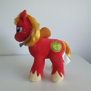 May include: A plush red pony toy with a yellow mane and tail. The toy has a green apple design on its side and tan hooves. It has a small, gray accessory attached to its mouth.