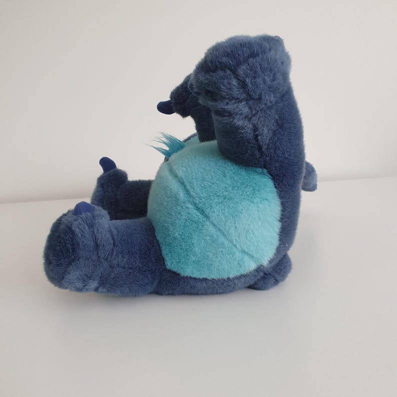 May include: A plush toy in the shape of a blue alien character, with a light blue belly and dark blue fur. The toy is lying on its back, with its arms and legs outstretched. The toy has large ears and a small tuft of hair.