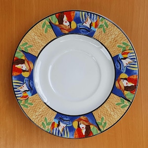 Könnte beinhalten: Eine weiße Untertasse aus Keramik mit einem blauen und gelben Rand. Der Rand zeigt ein sich wiederholendes Muster aus stilisierten Frauen mit Hüten. Die Untertasse ist mit einem farbenfrohen, abstrakten Design verziert.