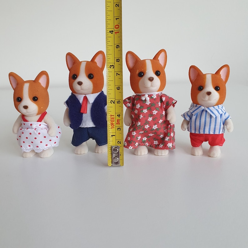 May include: Four toy corgi figures, each with a white and brown coat. One wears a red and white polka dot dress, another a navy suit with a red tie, a third a red floral dress, and the last a blue striped shirt and red shorts. A yellow ruler is next to the figures.