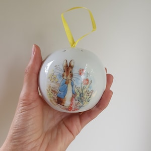 Puede incluir: Un adorno de cerámica blanca con una cinta amarilla. El adorno presenta una imagen impresa de Peter Rabbit con una chaqueta azul y zapatos rojos, de pie en un jardín con flores.
